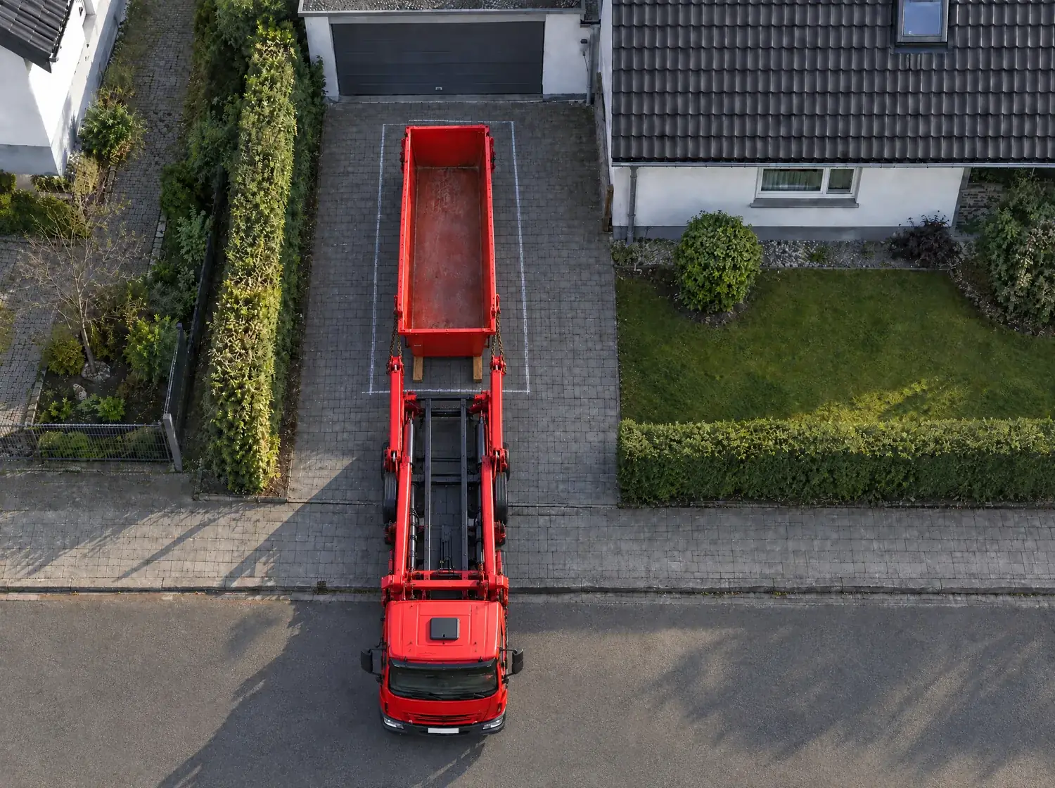 KI-Bild von oben: Ein roter Absetzkipper steht mit dem Führerhaus auf der Straße und reicht über den Gehweg in die Einfahrt. Dahinter hat er die lange rote Mulde vor der Garage abgesetzt, auf zwei Bohlen in einem Kreiderechteck.