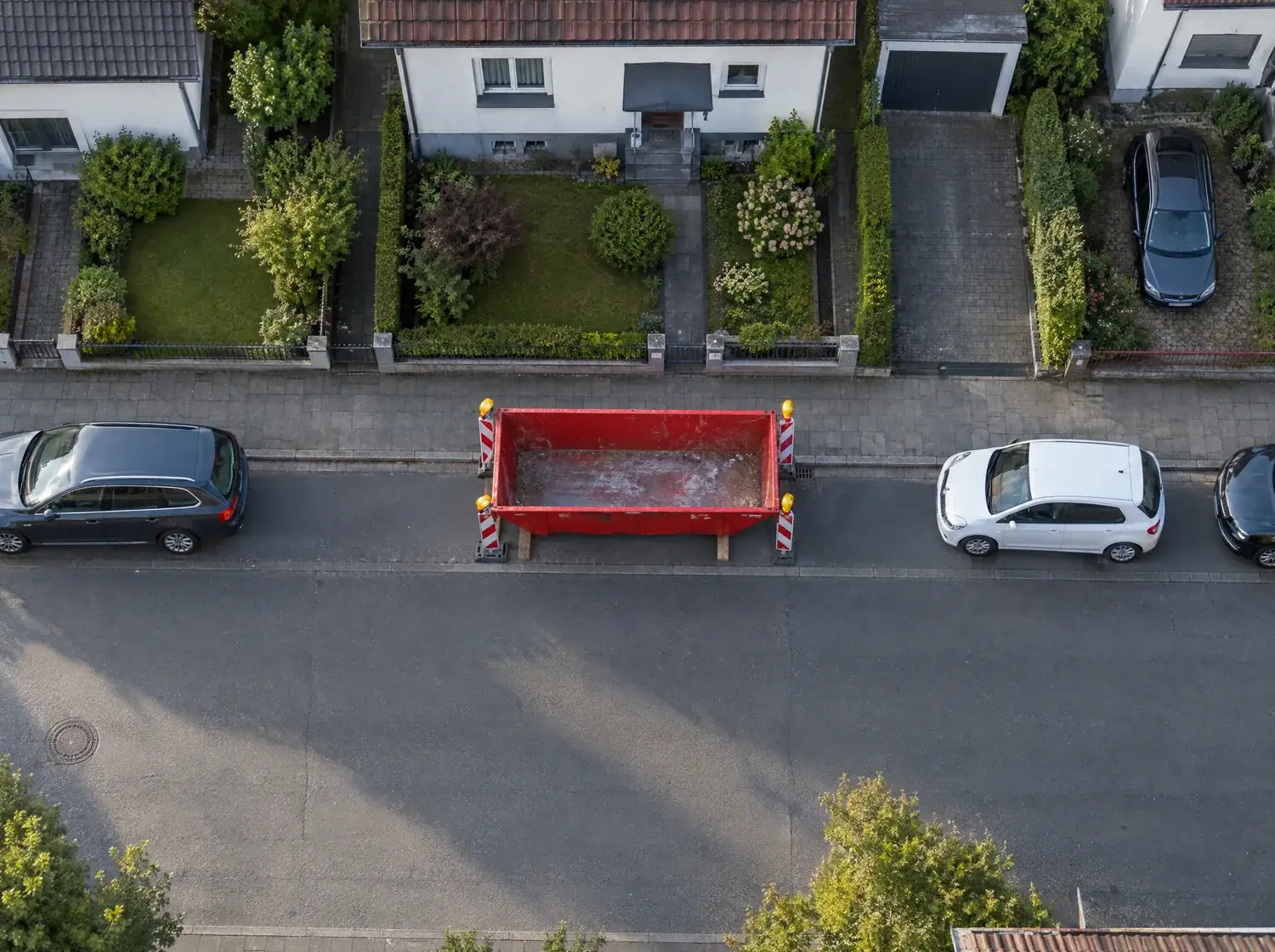 KI-Bild von oben: Rote Mulde in der Parkspur zwischen zwei Autos, auf zwei Bohlen, an beiden Enden eine rot-weiße Warnbake mit Licht. Gehweg und Fahrbahn bleiben frei.