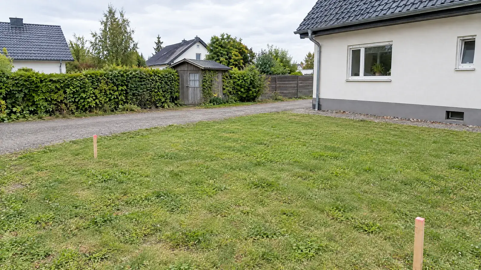 KI-Bild, vorher: Rasenfläche neben dem Haus, zwei Pflöcke markieren den Anbau